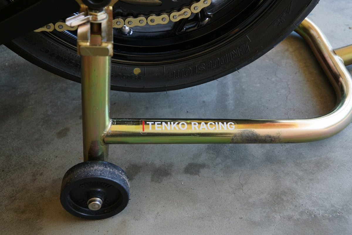 Gold motorcycle stand with 'Tenko Racing' branding supporting a motorcycle wheel on a concrete floor.